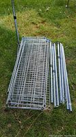 Stack of 6 steel wire shelving shelves on grass with 8 cylindrical metal poles beside them.