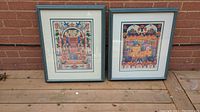 Two blue-framed lithographs by Michael Elkayam resting against a brick wall, showing detailed colorful designs with Jewish cultural themes.
