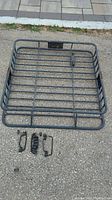 Top view of rectangular metal rooftop storage rack laid flat with mounting hardware arranged nearby on pavement.
