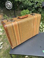 Photo of the vintage striped tweed suitcase placed on a black metal table, showing front, handle, and corner edges with visible leather trim and brass hardware.