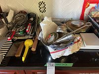 Photo of baking and cooking utensils on a black countertop including spatulas, strainers, tongs, measuring cups, a yellow citrus juicer, wooden rolling pin, metal funnel and a white food processor.