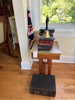 Photo showing small wooden table with stack of vintage books, brass hand bell, and British flag in corner.