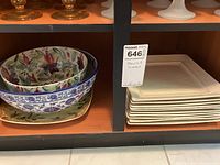 Shelf with stack of 6 cream-colored square stoneware plates on right and stack of 4 decorative bowls on left, including floral and blue/white patterned bowls.