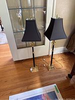 Pair of candlestick lamps shown side by side near a glass door on a wooden floor. Lamps have black shades and gold square bases with black vertical stems and gold accents.