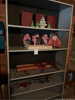 Shelving unit holding all Christmas decor items including ceramic trees, glass jars, fabric Santa figurine, and wooden sled.