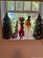 Front view of the lot arranged on a window ledge. Shows two decorated artificial trees, wooden reindeer, wooden tree, snowman figure, and undecorated tree.