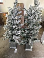 Two small artificial Christmas trees with snow flocking and white berry decorations, each on a gray textured stand. Background shows garage storage boxes.