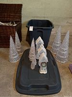 Full view of six cone-shaped translucent battery operated Christmas trees in various heights and three decorative white houses with snow-covered roofs and holiday wreaths, placed on a black plastic storage bin and floor.