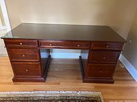 Large wooden desk with glass top and multiple drawers shown fully assembled on wooden floor.