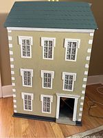 Front view of wooden doll house showing three floors, six windows with lace curtains, beige exterior, white trim, and blue roof.
