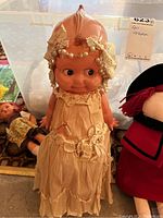 Vintage Kewpie doll wearing a cream dress with lace and pearl accents, standing upright with distinctive large head and big eyes.