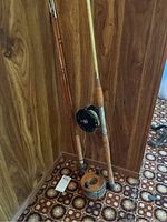 Two wooden fishing rods with reels leaning in a corner against wood panel walls and carpeted floor.