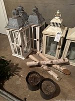 Four decorative lanterns with candles, birch logs, and two pots with soil on concrete floor