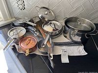 Full view of the set of pots and pans showing multiple stainless steel cookware pieces and one copper pot, all with lids and handles, resting on a black countertop.