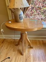 Oval blonde wood side table with central pedestal base shown near window on wooden floor with power cord visible.