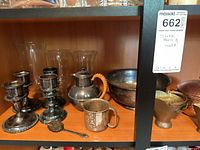 Shelf displaying silver bowl, two sets of candle holders, teapot with wicker handle, Mickey Mouse cup, and decorative spoon.