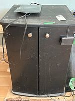 Front view of the black aquarium stand showing two doors with silver knobs, a dusty electric pad on top with cord attached.