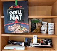 Photo showing Grillaholics grill mats, smoking chips in containers, pellet smoker tube, flat metal skewers, and Weber recipe binder on shelf.