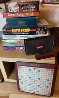 Photo of stacked board games including Lost The Game, Great Paper Jets, Fields of Assault Risk, Battleship, Suomipeli, Monopoly Deluxe Edition, Parcheesi Deluxe Edition, and Scrabble sets with carrying case