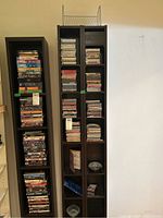 Two tall, dark shelving units filled with CDs and DVDs, first unit contains mixed CDs and DVDs with some visible titles, second unit contains mainly CDs and some other media items, wire CD rack placed atop second unit.