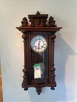 Front view of antique wooden pendulum wall clock hanging on wall, showing glass door, clock face with Roman numerals, and visible pendulum.
