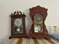 Two clocks displayed side by side on a shelf, left clock with wood and glass case, right clock with a plastic case designed to resemble wood with carved details and glass front.