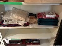 Shelf with stacks of assorted kitchen linens including napkins, tablecloths, tea towels and packaged coffee filters. Various colors and folded textile items, some in plastic packaging.