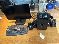 Full lot photo showing Acer monitor, Microsoft keyboard, Logitech speaker system with subwoofer and five speakers, along with cables on a wooden floor.