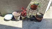 Photo showing 4 ceramic planter pots in various sizes and colors, 2 plastic planters filled partially with soil and plants, 2 plastic hanging baskets, and a metal plant stand on a concrete surface outside.