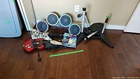 Photo of Guitar Hero and Rock Band instruments including RedOctane X-Plorer guitar controller, Rock Band drum set with pedal and stands, and various accessories and cables laid on the floor.