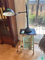 Full view of the black adjustable desk lamp standing on a distressed wooden footstool by a window. Shows the lamp, footstool, and the state of paint wear on the stool.