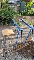 Two blue metal folding sawhorses and a small wooden press stand with metal legs shown outdoors in garden setting.