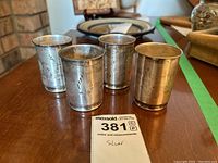 Four small silver cups arranged together showing tarnishing and engraved decorative pattern.