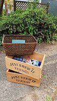 Two wooden boxes stacked, both labeled 'JUST BREW IT VERNON' with a large woven basket behind them containing various items.