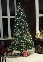 Decorated Christmas tree standing near large windows, adorned with warm white lights, ornaments, and gift boxes beneath.