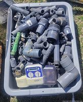 Top view of gray plastic hose fittings of various sizes and types, a green connector, and an Orbit sprinkler timer in a gray bin outdoors on grass.