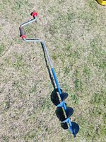 Full view of the 7 1/2 inch manual ice auger showing the crank handle with red grips and spiral blade on grass background.