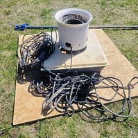 White cylindrical fountain nozzle housing atop square base plate on plywood, with coiled black power cord and braided rope beside it