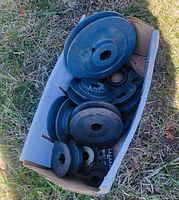 Box containing assorted steel pulley wheels of various sizes and types, resting on grass outdoors.