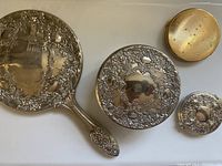 Top down image showing all 4 items: handheld mirror, trinket box with silver-plate lid, silver-plate mirror compact, and additional gold-tone powder compact.
