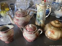 Seven-piece tea set including four teapots, creamer, and sugar bowl displayed on a table with various glassware in the background