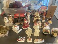 Wide view of all Christmas figurines, candles, vintage Coca-Cola ornament, and snowman salt and pepper shakers laid out on surface.