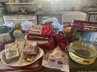 Photo showing an assortment of holiday-themed kitchen and serving items including red teapot, napkins, trays, and glassware boxes
