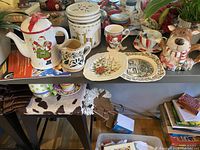 Overview of holiday ceramic tea set items on table including teapot, cookie jar, mugs, plates, and tea cups