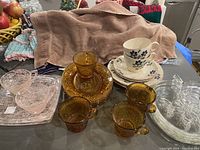 Full lot overview showing all plates, cups, and trays including Hazel Atlas Pink Drizzle clear glass tray with pink drizzle pattern, Indiana Glass Tiara Amber cups and plates, clear apple glass plates, and Mikasa Country Classics cups and plates.