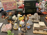 Wide shot showing grouped vintage figurines including boxed Shoe Box Bears, Dept 56 Snow Babies in green boxes, Hello Kitty figure, animals, and small decorative building models.