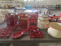 Red kitchen decor items including cookbook stand, candle holder, spoon rest, napkin holder, trivet, black iron plate holder with apples, and cream/red enamel cookware consisting of pot and double boiler.