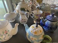 Lot of five teapots with various floral and colored designs, matching cream and sugar bowl, and one teacup with a teaspoon on a dark surface with window light.