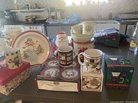Full lot view showing large snowman serving plate, hard plastic holly plates and mugs, vintage boxed mugs, Debbie Mumm boxed plate set, Santa cookie jar, poinsettia tin, and napkins.