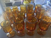 Front view of 15 amber Indiana Glass goblets showing different sizes for water and wine glasses, warm gold tone and vertical fluted pattern on bowls, round stems, round bases.